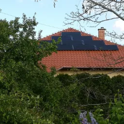 Panele słoneczne na czerwonym dachu domu jednorodzinnego, widoczne zza zieleni drzew i krzewów w słoneczny dzień.