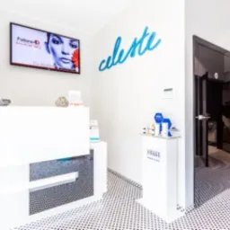 Jasne wnętrze salonu kosmetycznego z białą recepcją z czarnymi elementami, telewizorem z reklamą zabiegu kosmetycznego na ścianie oraz dekoracyjnym napisem 'celeste' nad recepcją. Widoczne drzwi...