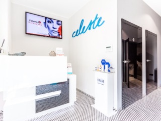 Jasne wnętrze salonu kosmetycznego z białą recepcją z czarnymi elementami, telewizorem z reklamą zabiegu kosmetycznego na ścianie oraz dekoracyjnym napisem 'celeste' nad recepcją. Widoczne drzwi...