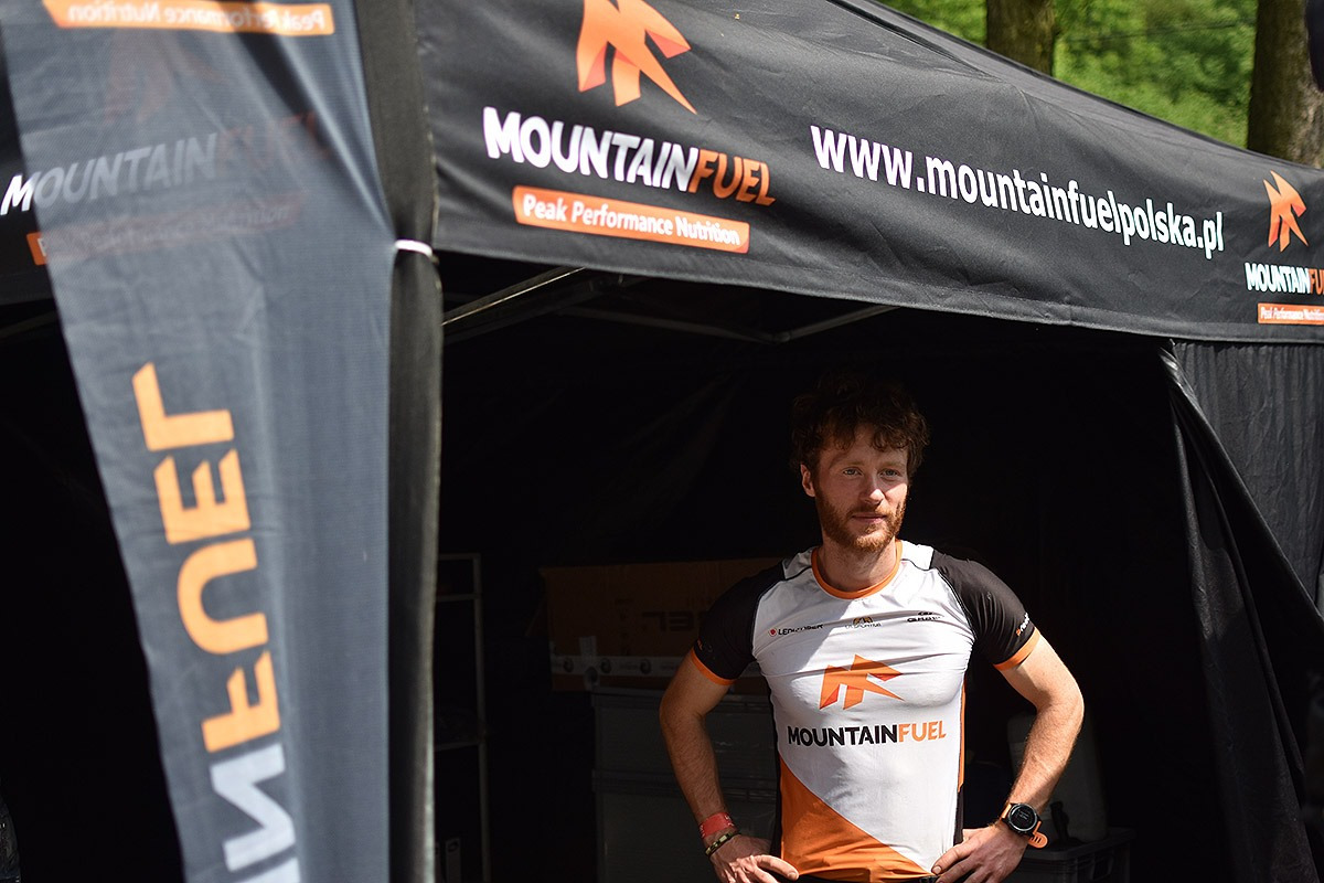 Mężczyzna w sportowej koszulce z logo sponsora stoi pod namiotem firmy produkującej odżywki dla sportowców podczas zawodów biegowych.