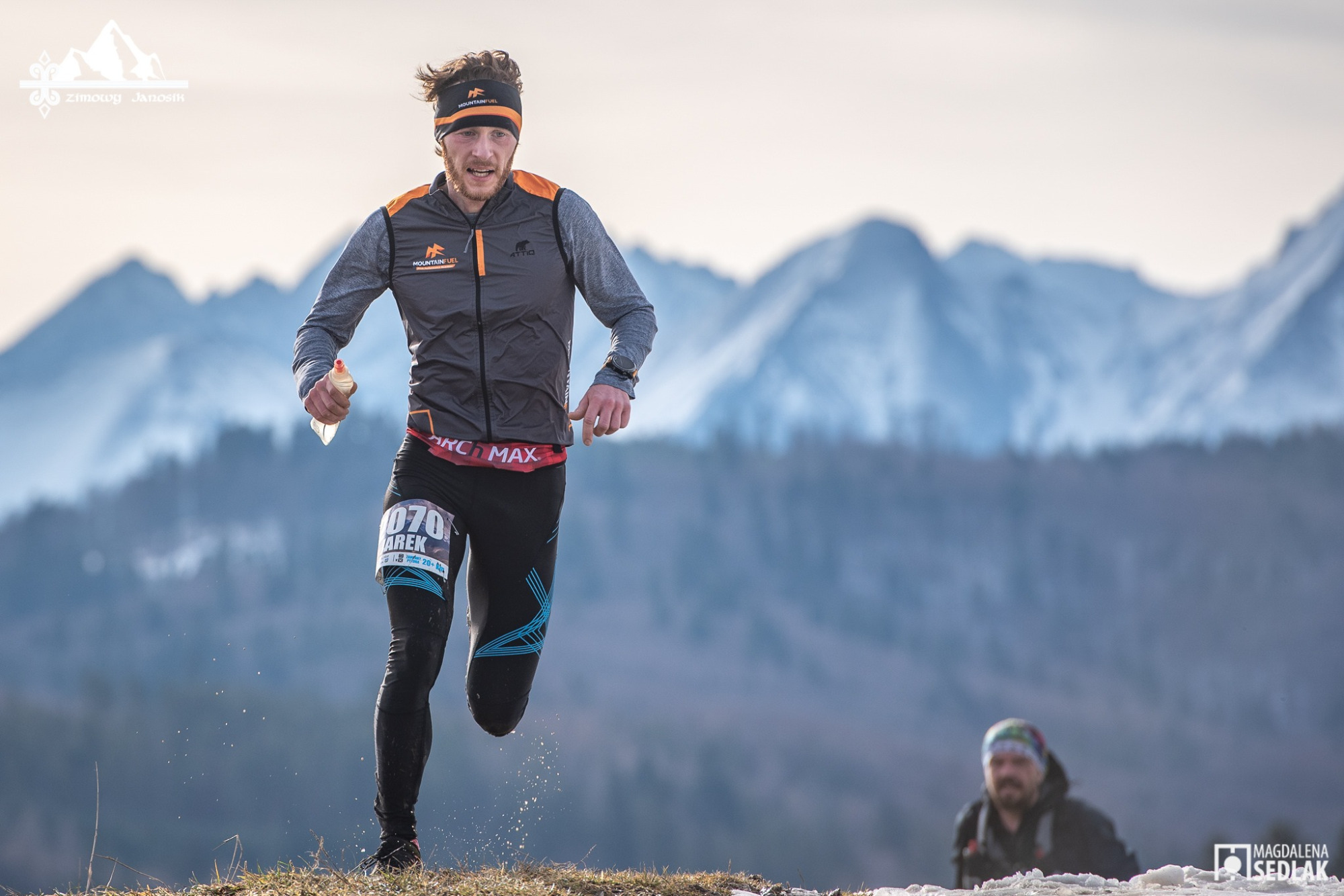 Biegacz górski w trakcie zawodów, widoczny numer startowy 070, w tle ośnieżone szczyty górskie i inny zawodnik.