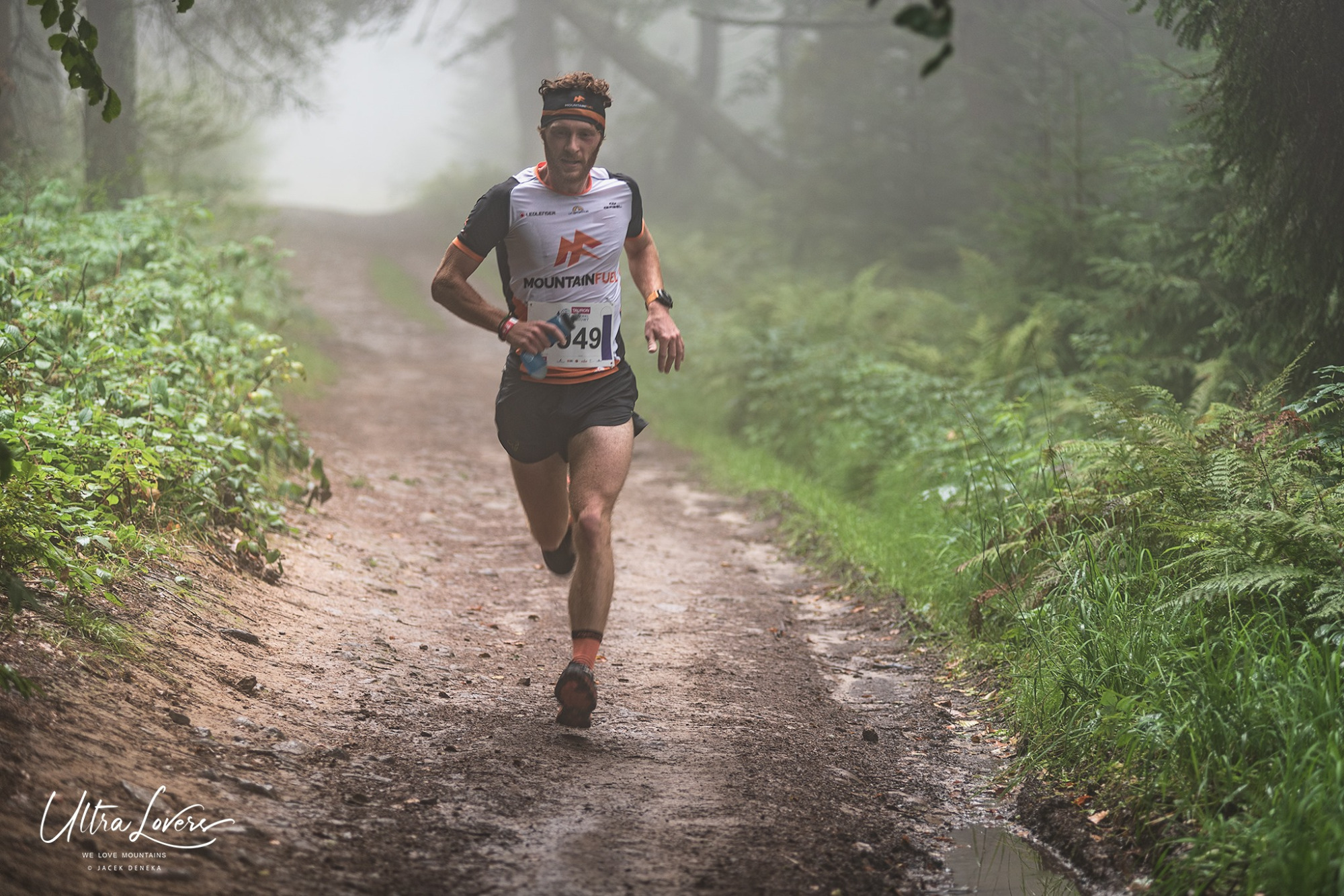 Mężczyzna w sportowym stroju biegnie leśną ścieżką w lekkiej mgle, widoczny numer startowy 349, zarośla po bokach.