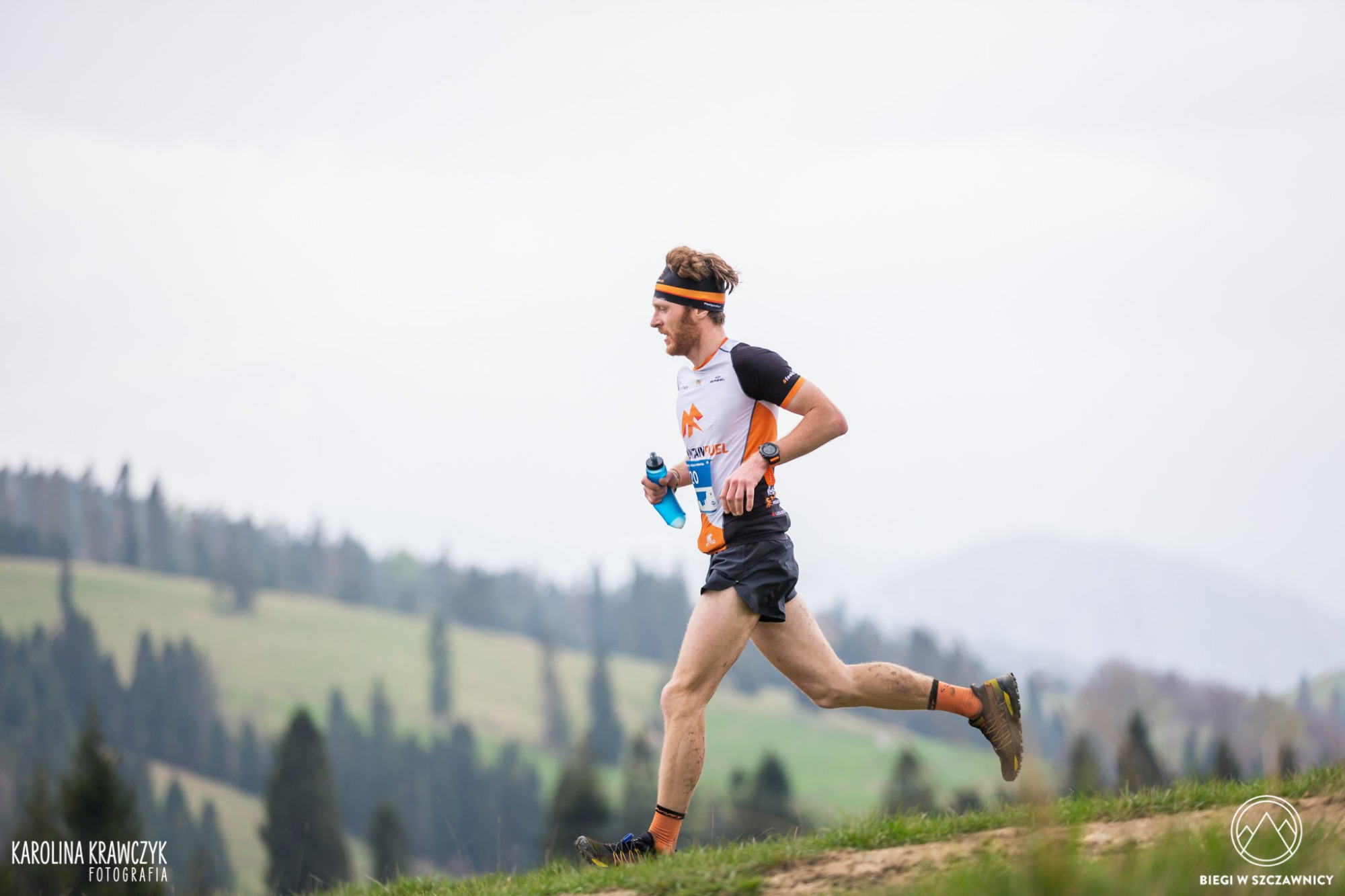 Mężczyzna biegnący w górach podczas zawodów, widoczny numer startowy i butelka z wodą, w tle zalesione wzgórza.