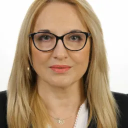 Portret uśmiechniętej kobiety w okularach, z blond włosami i delikatną biżuterią, reprezentującej firmę ubezpieczeniową we Wrocławiu.