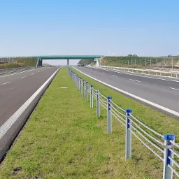 Nowa dwupasmowa autostrada z wiaduktem w tle, zabezpieczona metalowymi słupkami połączonymi linami, widok w słoneczny dzień.