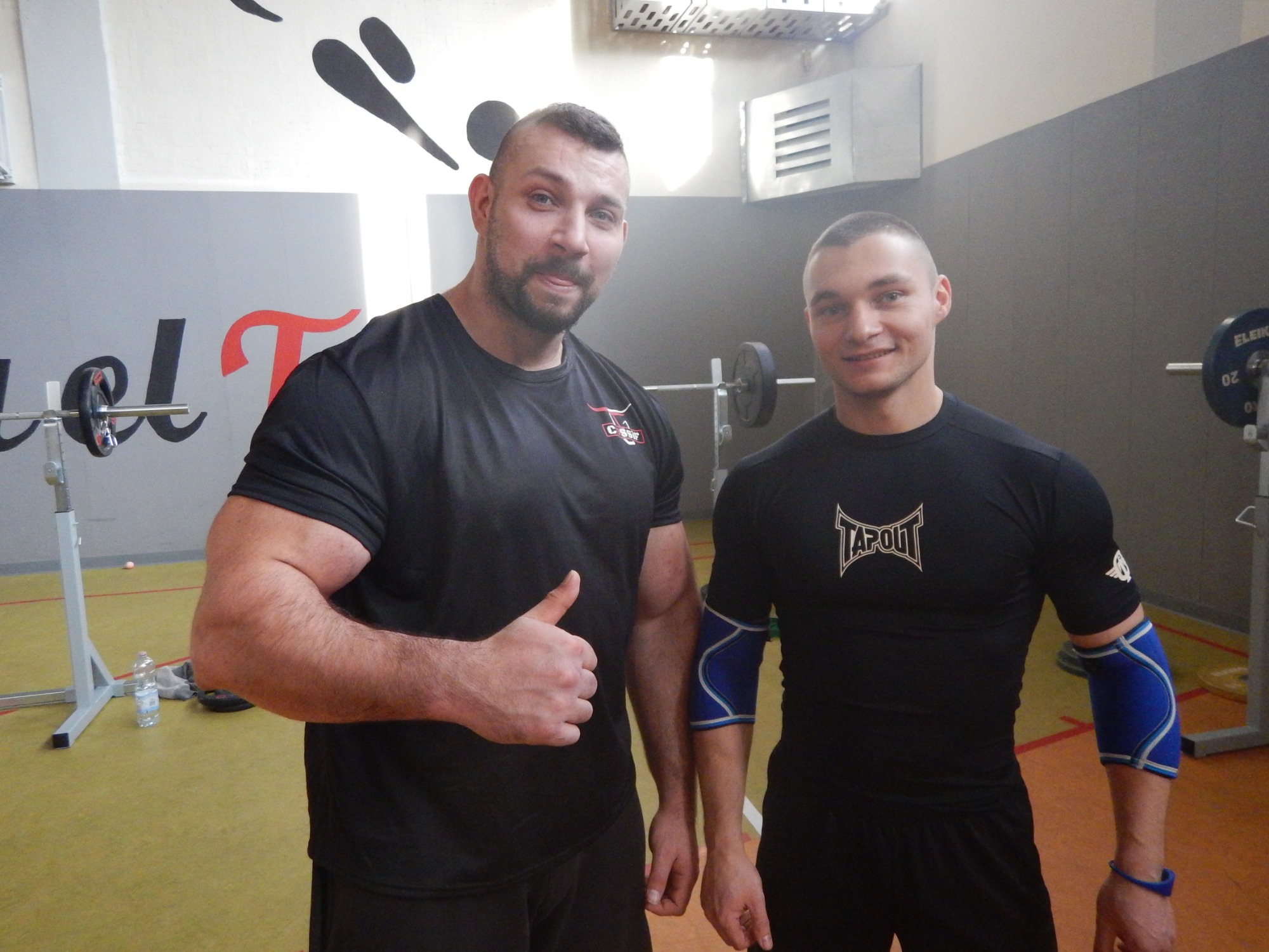 Dwaj mężczyźni w czarnych koszulkach pozują w siłowni, jeden z nich, muskularny, pokazuje kciuk w górę, w tle sztanga i sprzęt do ćwiczeń.