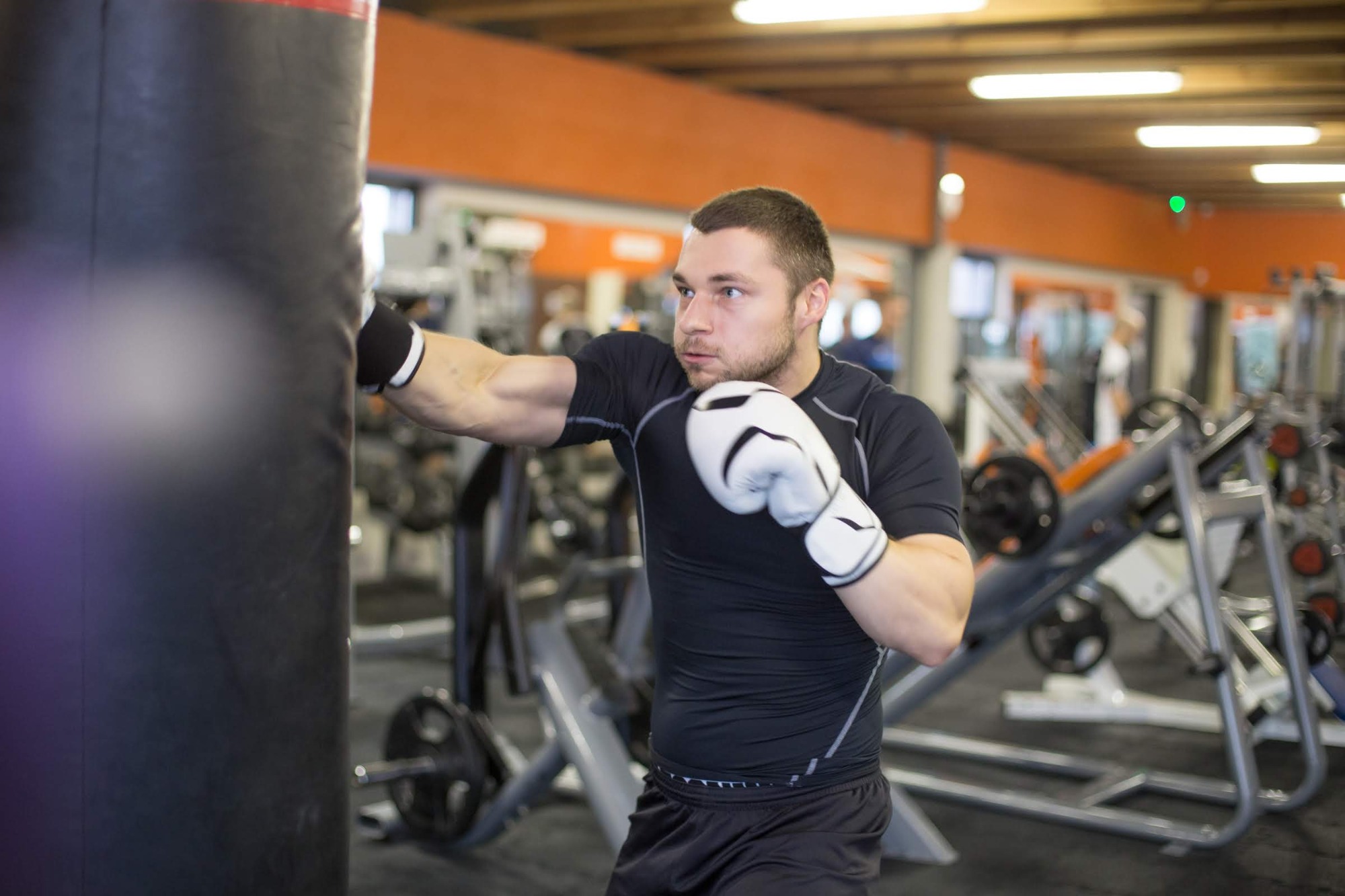 Mężczyzna w czarnym stroju i białych rękawicach bokserskich uderza w czarny worek treningowy w siłowni, w tle widoczne maszyny do ćwiczeń.