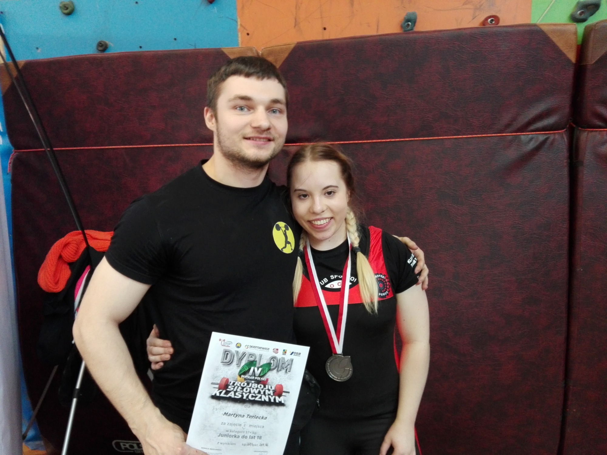 Uśmiechnięta para pozuje na tle ściany wspinaczkowej i maty; kobieta z medalem na szyi i dyplomem za trójbój siłowy w rękach mężczyzny.