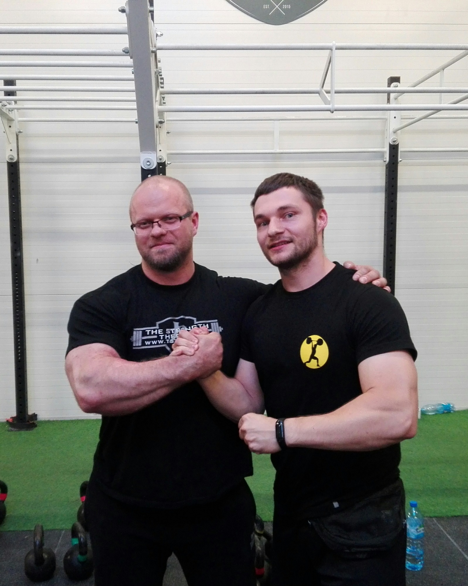 Dwóch mężczyzn o muskularnej budowie ciała, ubranych w czarne koszulki z logo, ściska sobie dłonie w siłowni, w tle widoczne kettlebells i sprzęt do ćwiczeń.