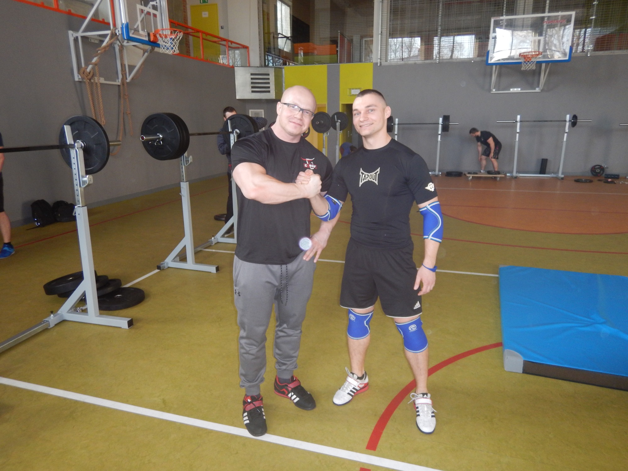 Dwóch mężczyzn w strojach sportowych w siłowni, z uśmiechem ściskają sobie dłonie, w tle sprzęt do ćwiczeń siłowych i koszykówki.