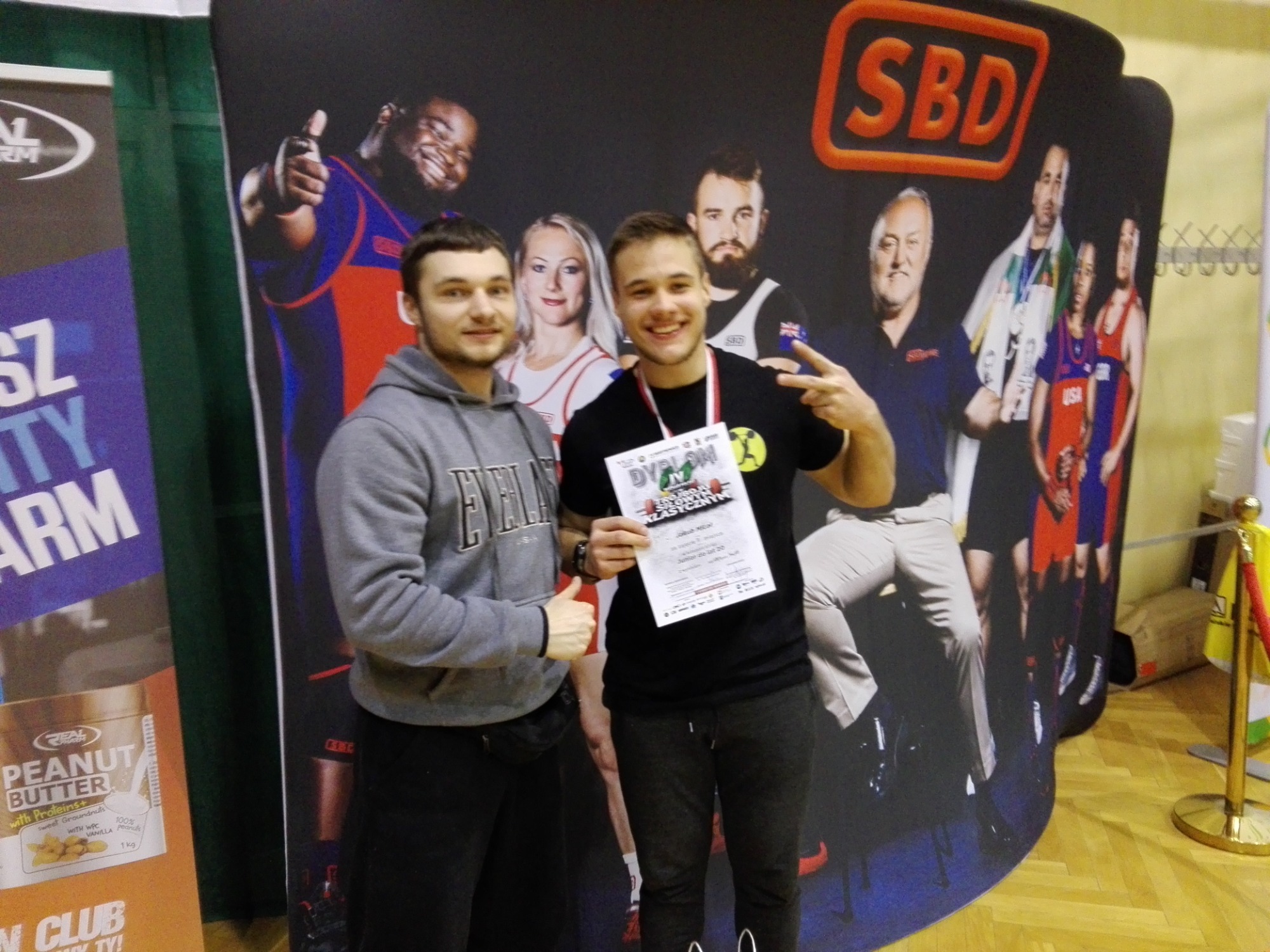 Dwóch młodych mężczyzn pozuje z dyplomem za IV miejsce w zawodach sportowych na tle baneru reklamowego z wizerunkami sportowców i logo SBD, obok widoczny baner z reklamą masła orzechowego.