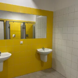 Wnętrze publicznej toalety z żółto-białymi płytkami, dwoma umywalkami na postumentach i dużym lustrem, widoczne boksy toaletowe w odbiciu.