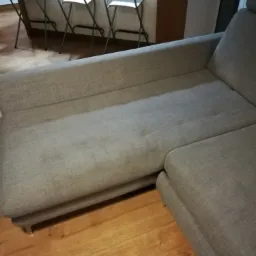 Szara, narożna kanapa z widocznymi plamami na tapicerce, ustawiona na drewnianej podłodze, w tle krzesła barowe.