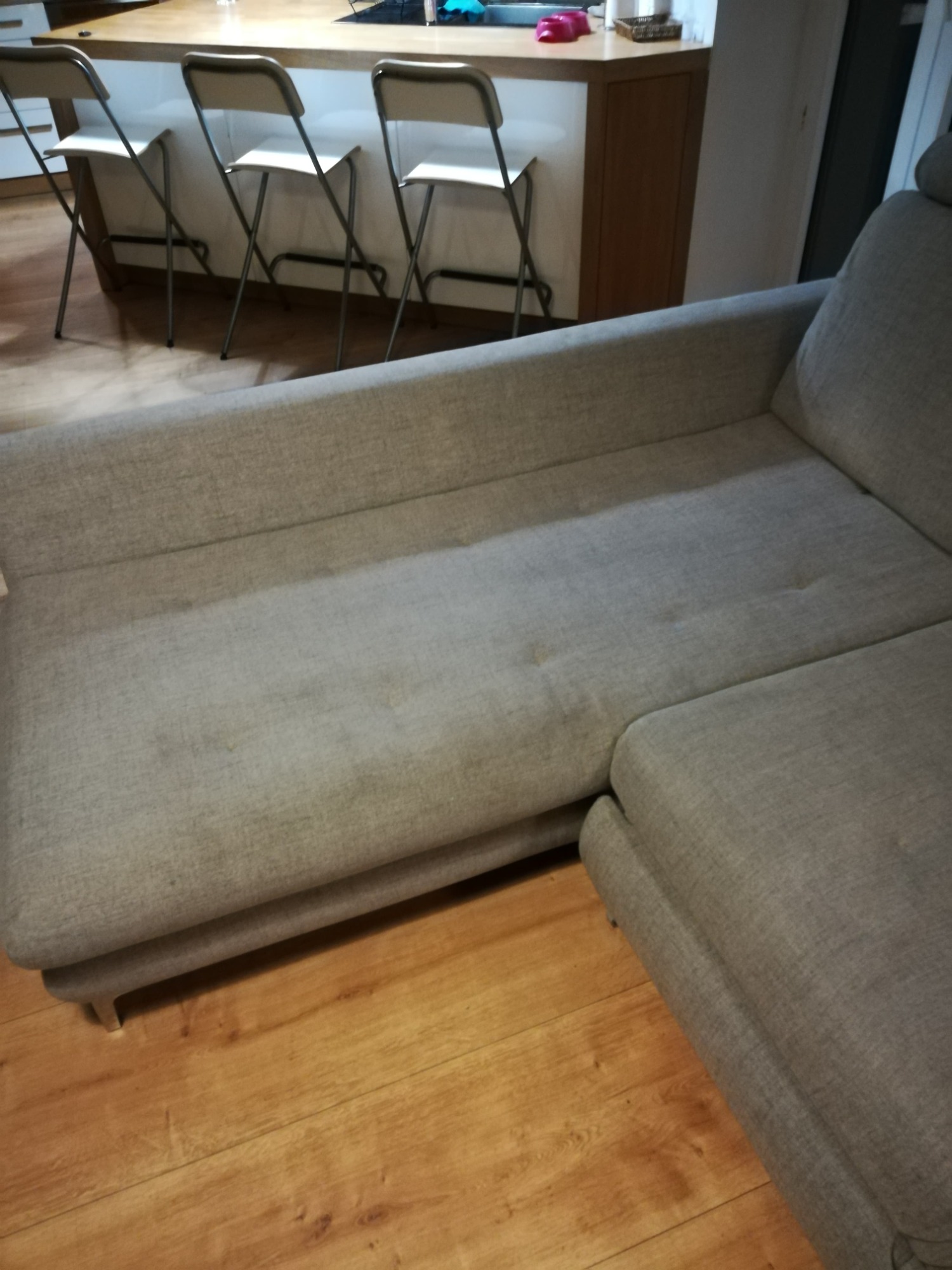 Szara, narożna kanapa z widocznymi plamami na tapicerce, ustawiona na drewnianej podłodze, w tle krzesła barowe.
