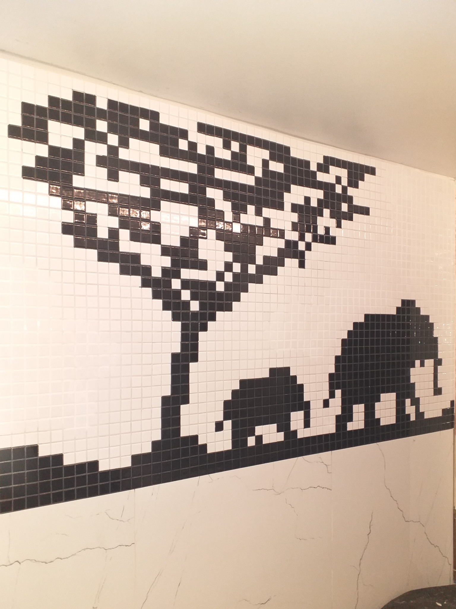 Mozaika łazienkowa przedstawiająca afrykański krajobraz z drzewem i słoniami, wykonana z czarno-białych kwadratowych płytek, z widocznymi pęknięciami na dolnej części ściany.