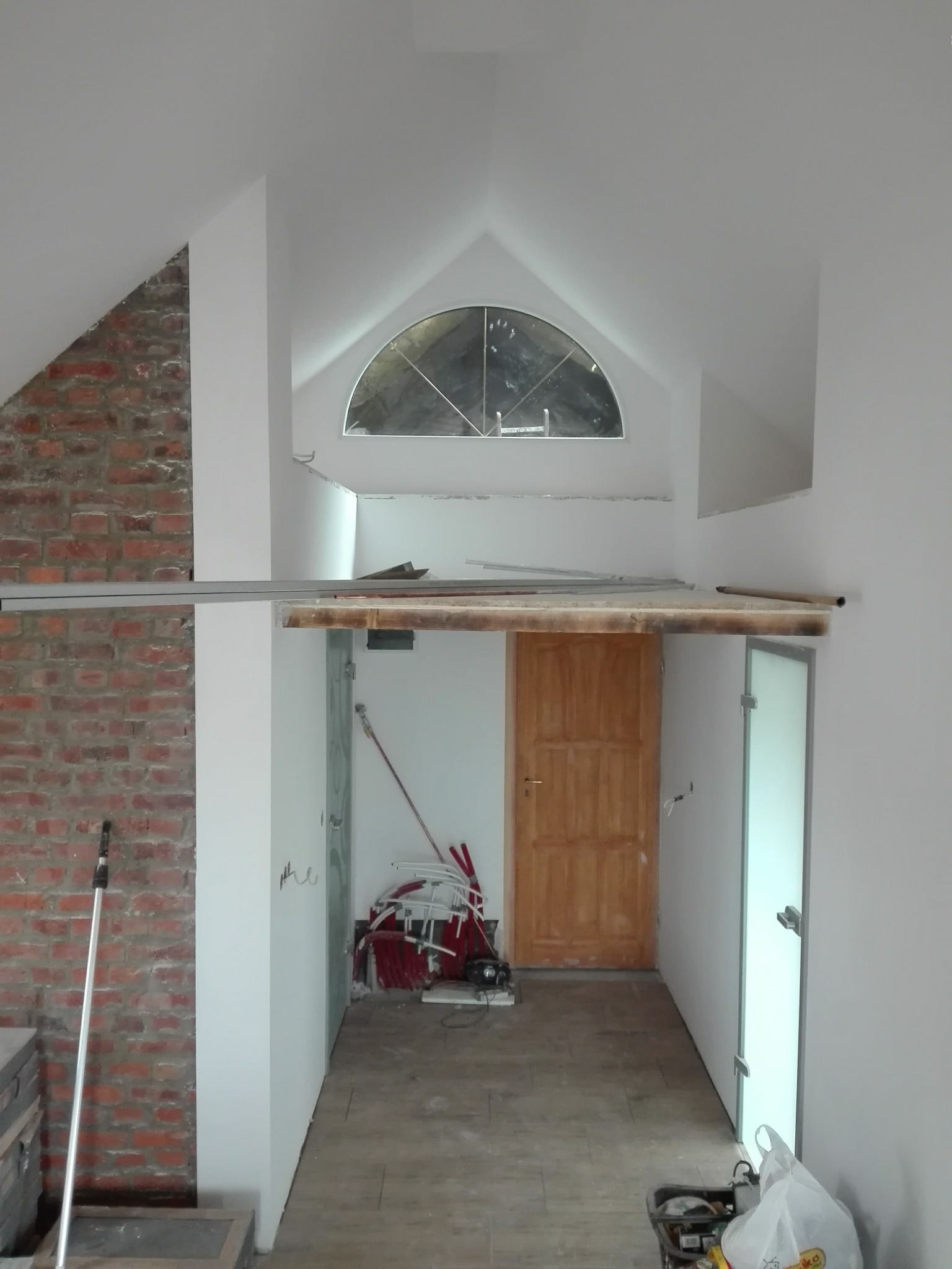 Wnętrze poddasza w trakcie prac wykończeniowych: widoczna ceglana ściana, okno w kształcie półkola, drewniane drzwi i narzędzia budowlane.