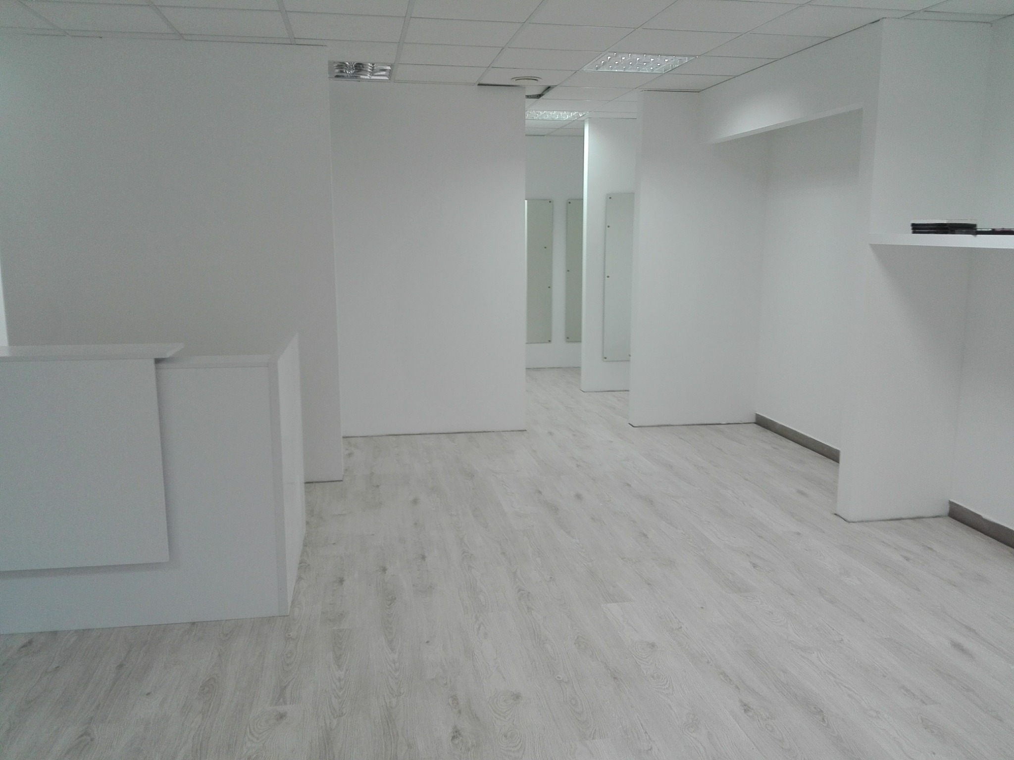 Wnętrze biura w odcieniach bieli z panelami podłogowymi imitującymi jasne drewno, widok na korytarz z lustrami, oświetlenie sufitowe rastrowe.