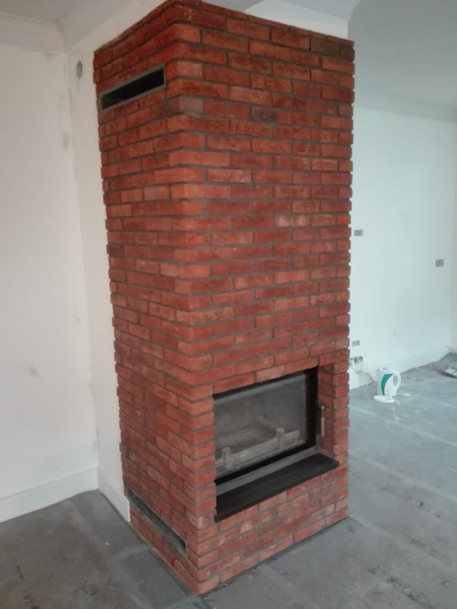 Murowany kominek z czerwonej cegły w trakcie wykończenia, widoczne surowe ściany i podłoga, czarny wkład kominkowy, otwór wentylacyjny u góry.