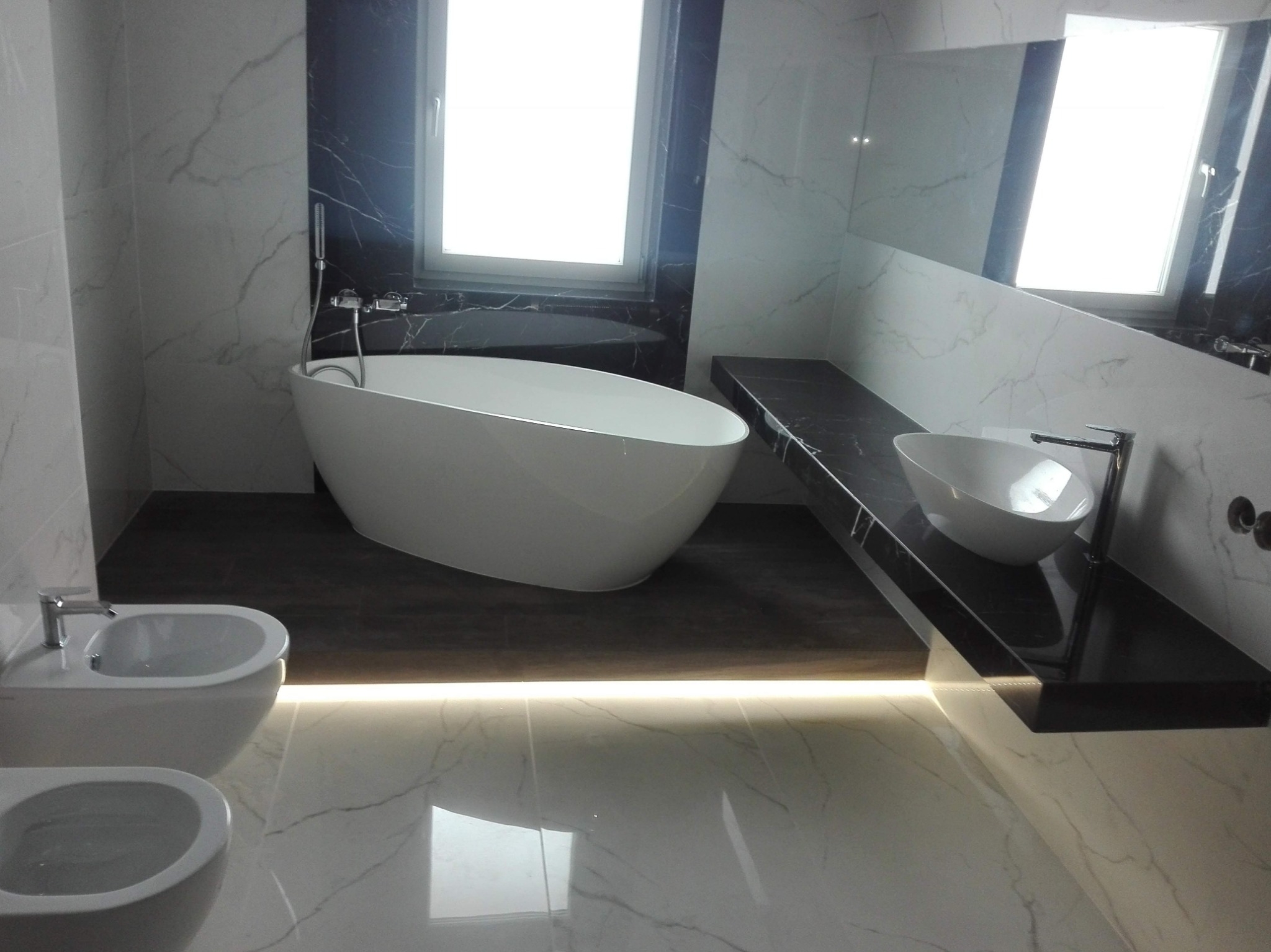 Elegancka łazienka z wolnostojącą białą wanną, umywalką nablatową i bidetem, wykończona marmurem z czarnymi akcentami i podświetleniem LED.