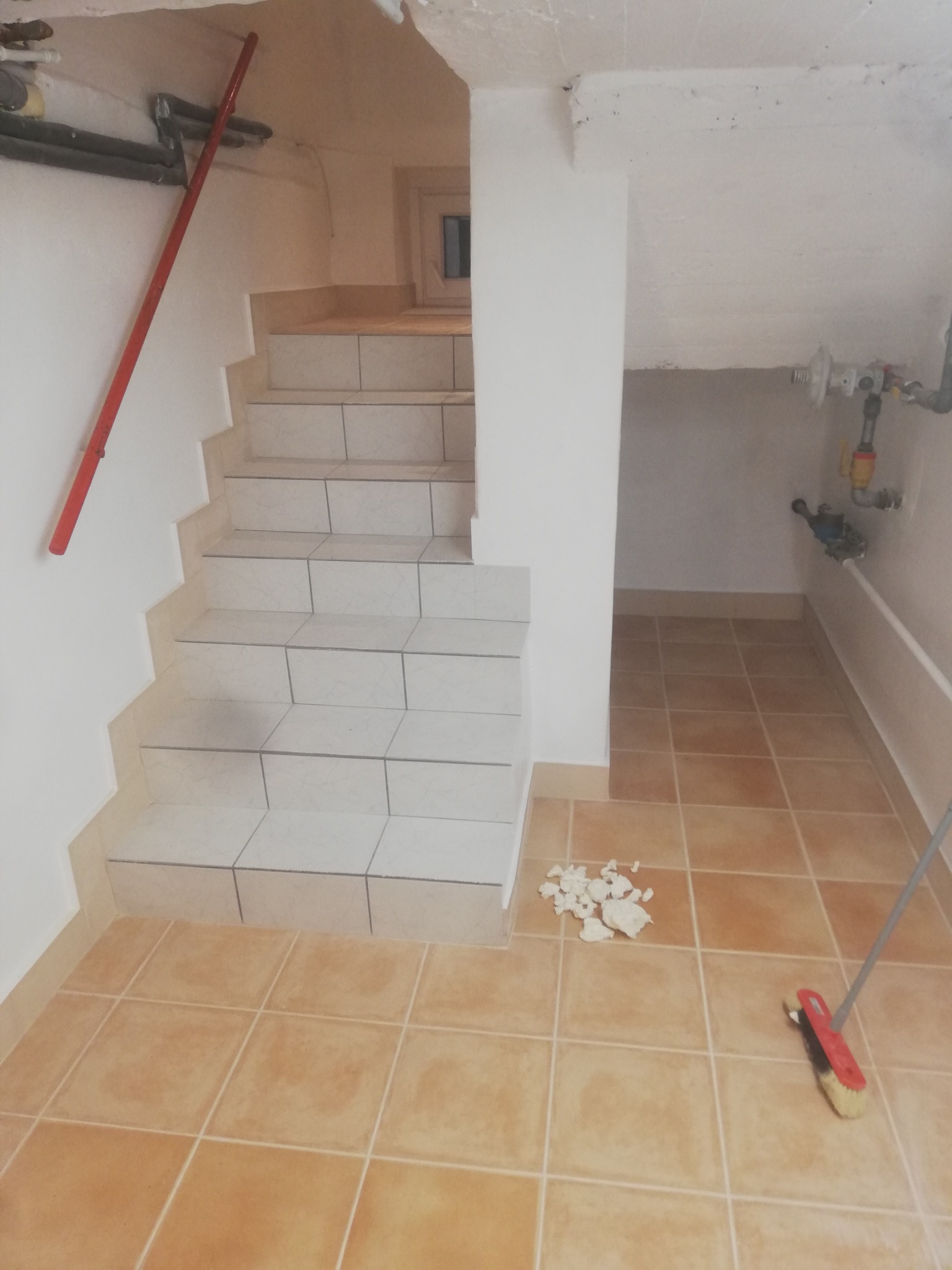 Klatka schodowa w piwnicy, wyłożona jasnymi płytkami, z widocznymi śladami po remoncie i stojącą szczotką do zamiatania.