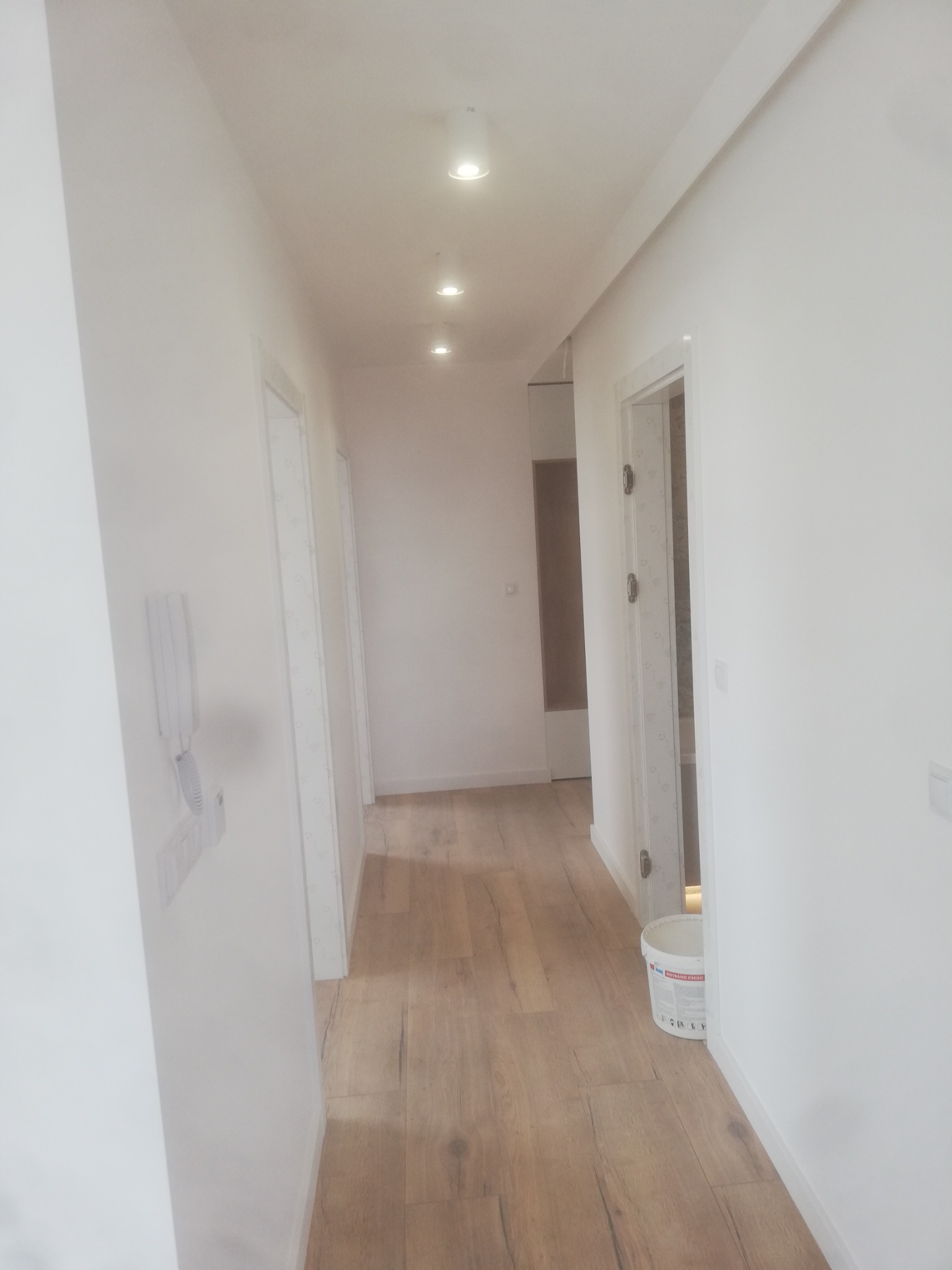 Wykończony korytarz w Bielsko-Białej z jasnymi panelami podłogowymi, białymi ścianami i sufitowym oświetleniem punktowym. W rogu widoczne wiadro po farbie.