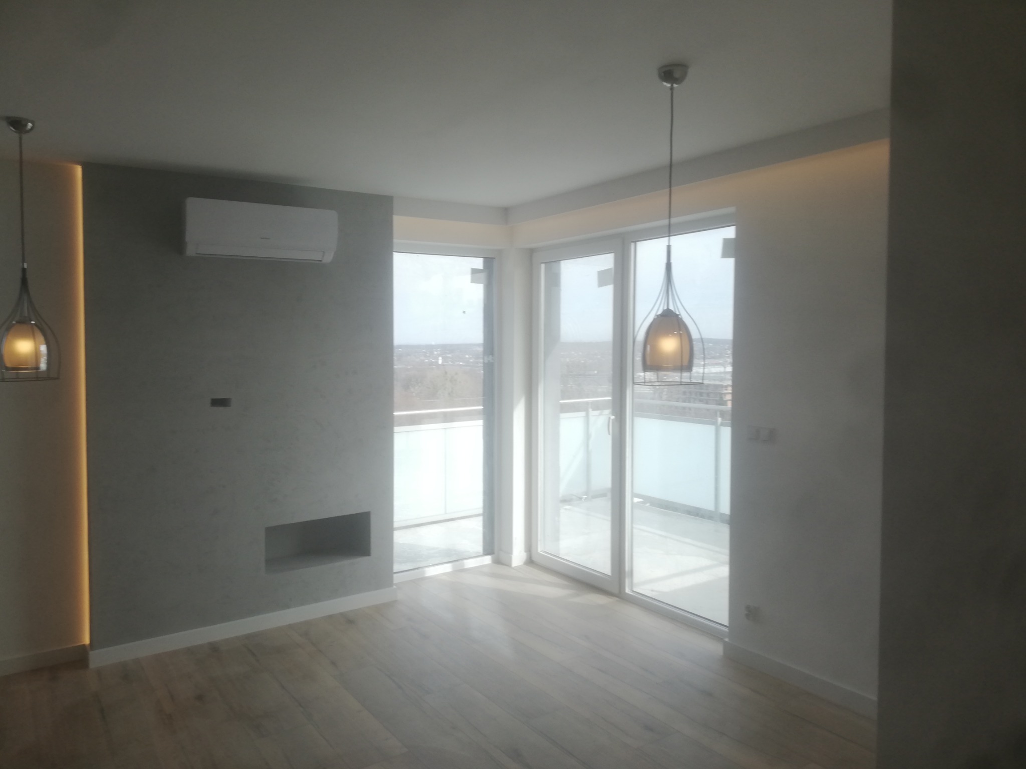 Pusty, jasny pokój z drewnianą podłogą, balkonem z przeszkloną balustradą, klimatyzacją na szarej ścianie z wnęką i wiszącymi lampami w metalowych kloszach.