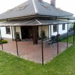 Zadaszony taras przy domu jednorodzinnym z metalową konstrukcją i przezroczystym dachem, na tarasie stół i krzesła, w tle budynek z oknami i kwiatami.