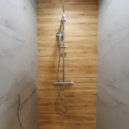 Nowoczesny prysznic z deszczownicą, wykończony białymi płytkami imitującymi marmur i drewnopodobnymi panelami ściennymi w odcieniach brązu.