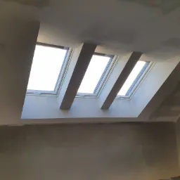 Trzy okna dachowe w surowym stanie, widoczne elementy konstrukcyjne i ślady szpachlowania na ścianach poddasza przed malowaniem.