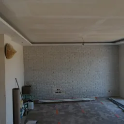 Pokój w trakcie prac wykończeniowych: ściana z dekoracyjnej cegły, częściowo wykończony sufit podwieszany z oświetleniem, widoczne przewody elektryczne, materiały budowlane i narzędzia.