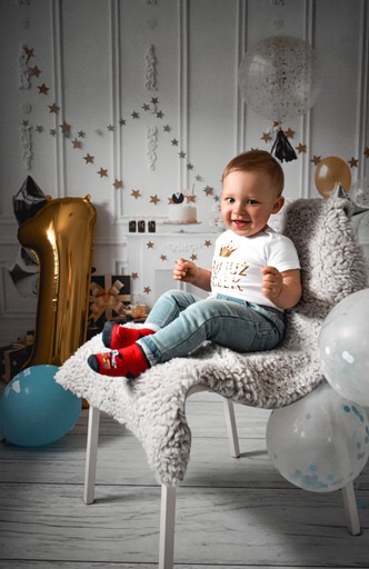 Uśmiechnięty roczny chłopiec w czerwonych skarpetkach siedzi na krześle z futrzanym siedziskiem, otoczony balonami i dekoracjami urodzinowymi w studio fotograficznym.
