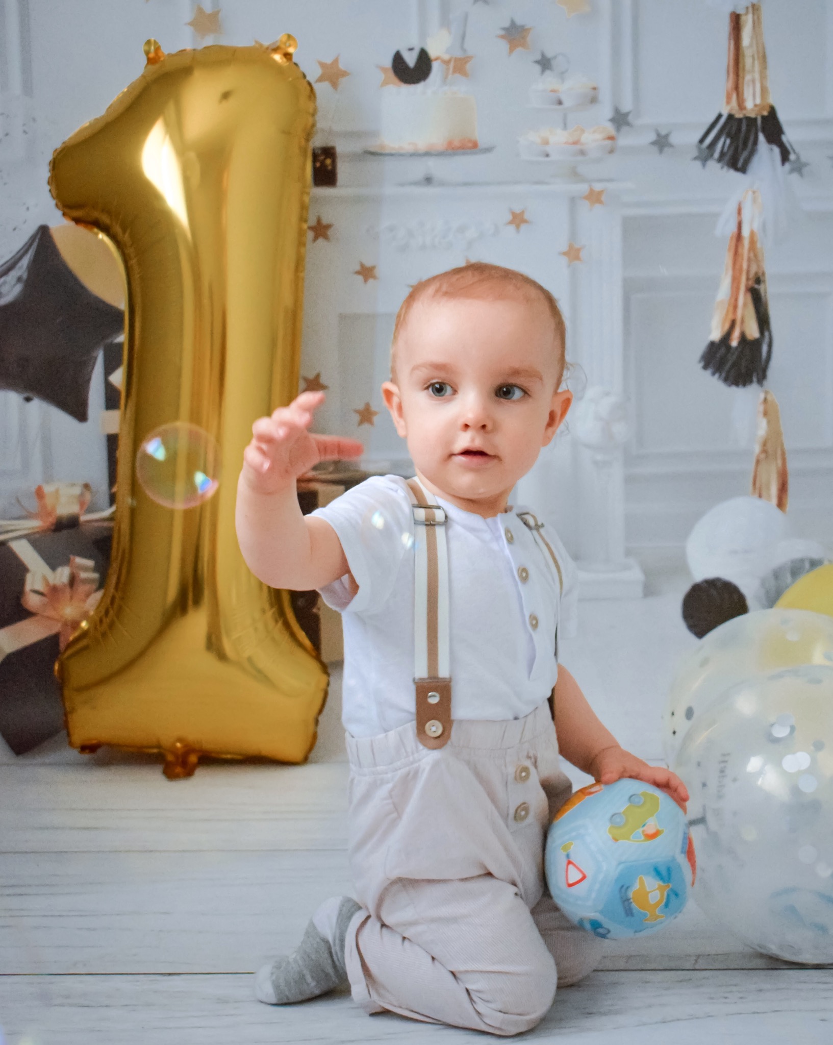 Roczne dziecko w białej koszuli i spodniach na szelkach, klęczy na podłodze z piłką w ręku, obok złoty balon w kształcie cyfry 1 i bańka mydlana, w tle dekoracje urodzinowe i tort.