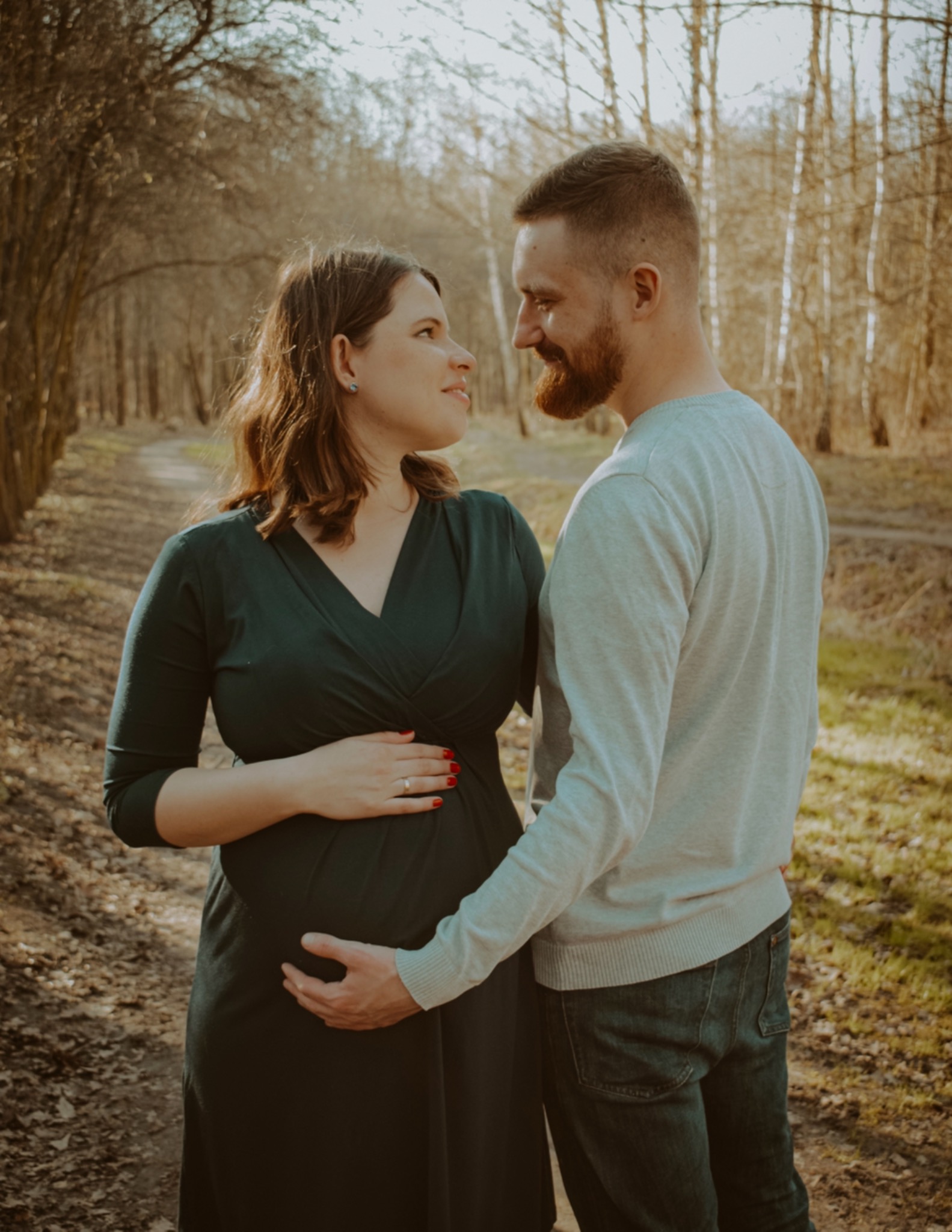 Przyszła mama w ciemnozielonej sukience, trzymająca dłoń na brzuchu, spogląda z uśmiechem na partnera z brodą, stojącego obok na leśnej ścieżce w słoneczny dzień.