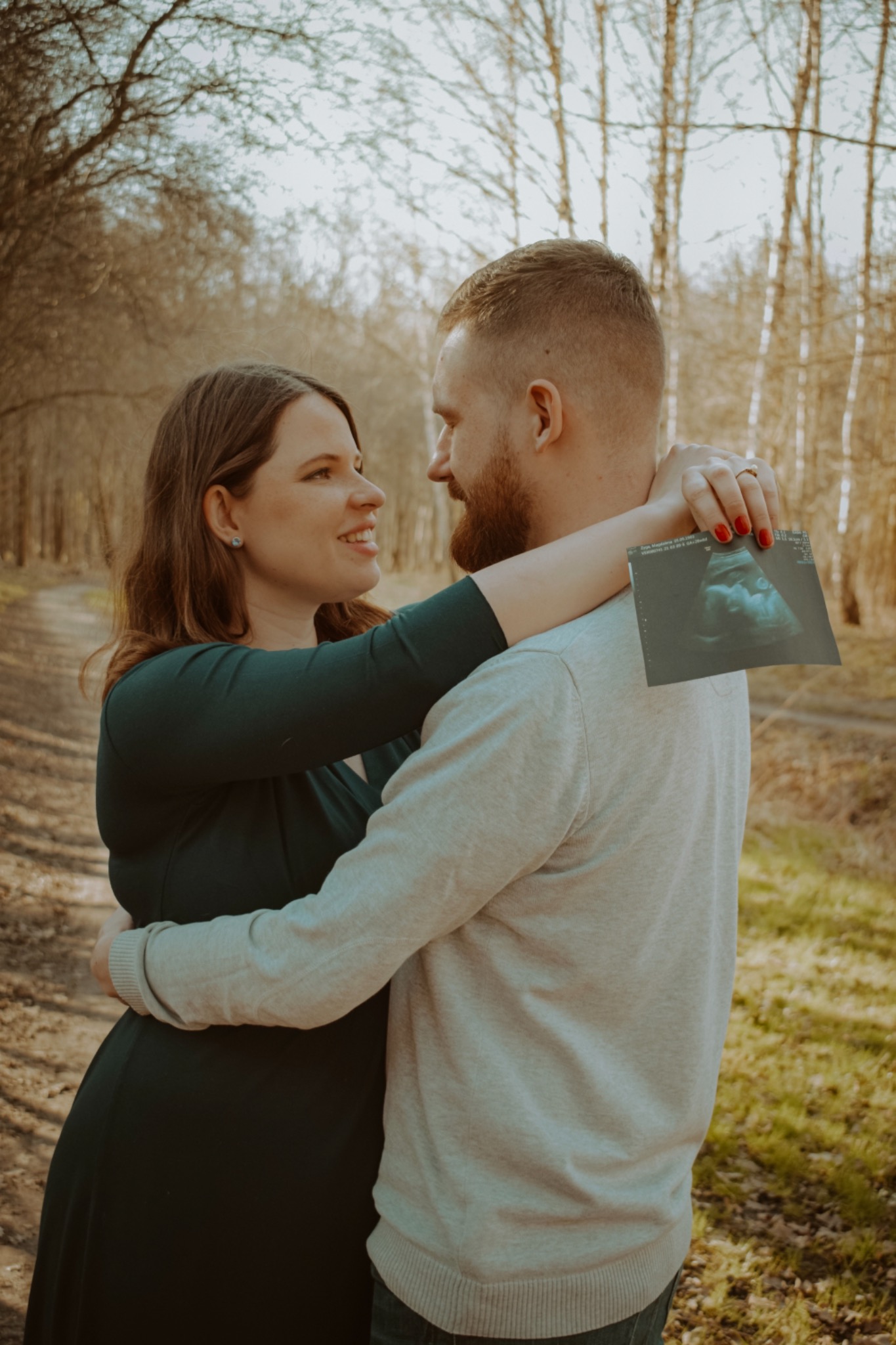 Zakochana para we Wrocławiu, kobieta w zielonej sukience ciążowej obejmuje mężczyznę, trzymając w dłoni zdjęcie USG dziecka na tle leśnej ścieżki.
