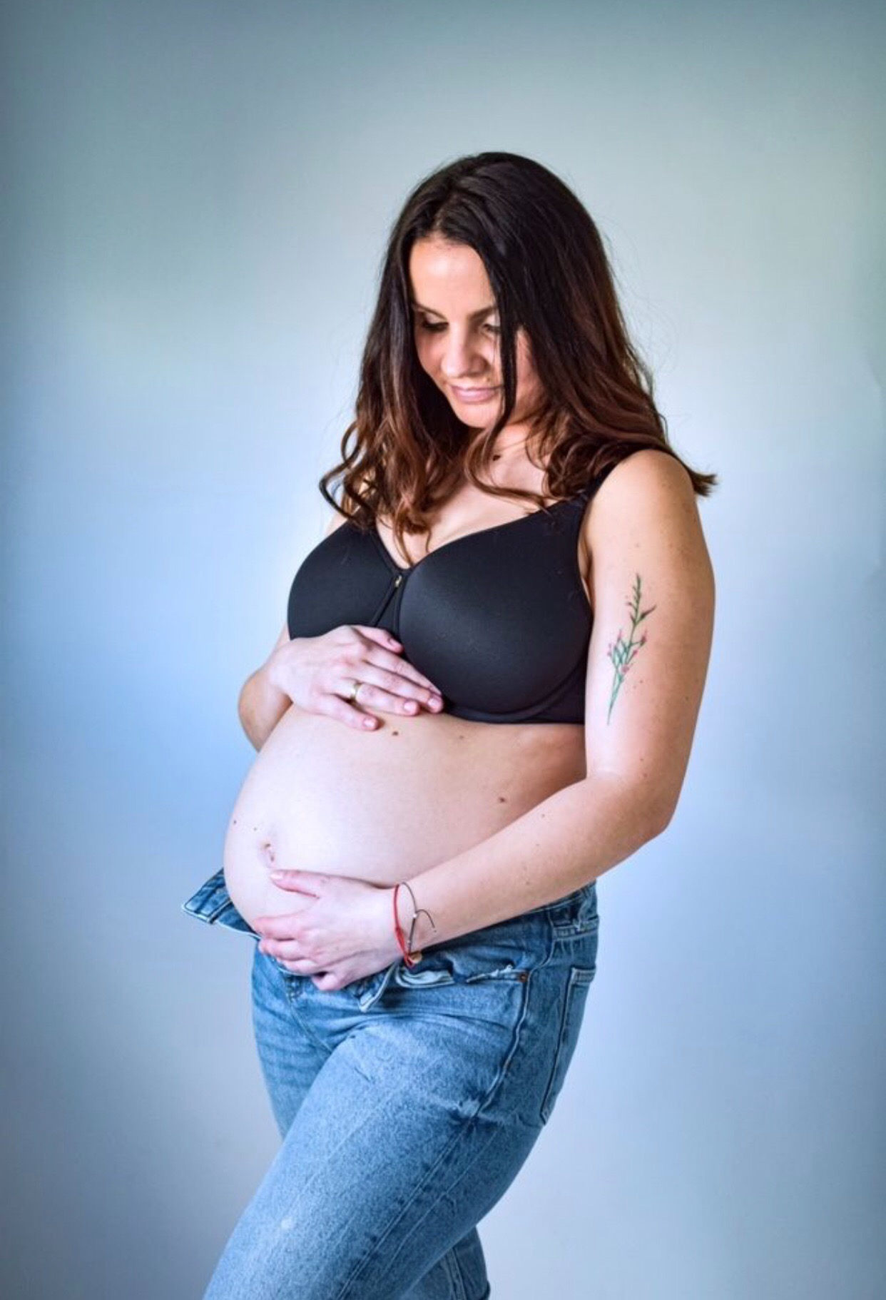 Kobieta w ciąży, ubrana w dżinsy i czarny stanik, delikatnie trzyma brzuch dłońmi, spoglądając w dół; widoczny tatuaż roślinny na ramieniu.