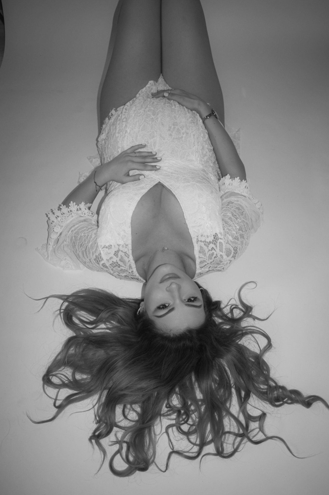 Czarno-białe zdjęcie kobiety w ciąży leżącej na plecach, ubranej w koronkową sukienkę, z rękami na brzuchu i rozłożonymi włosami na białym tle.