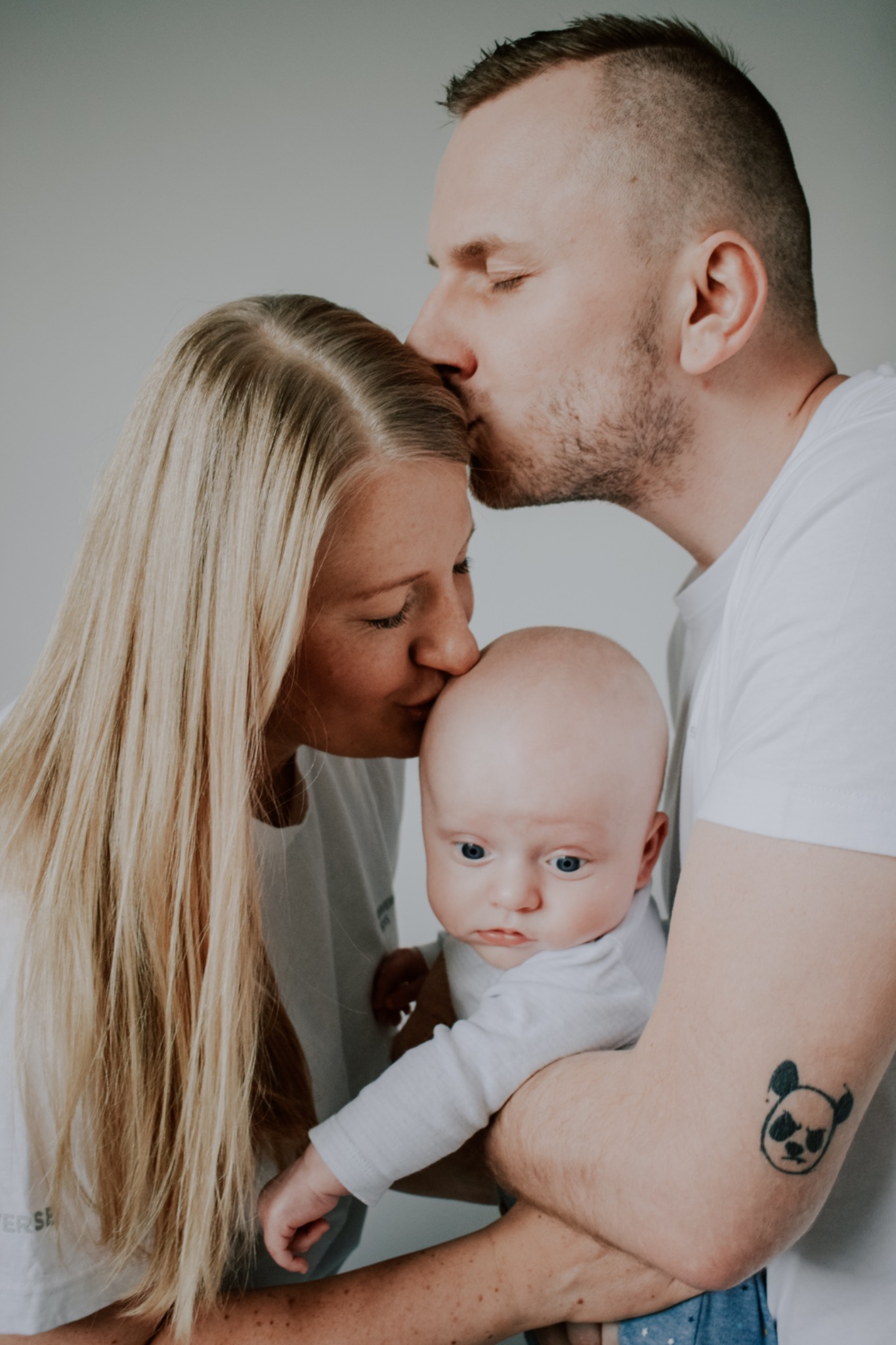 Rodzice całują niemowlę w głowę, tata ma tatuaż z pandą na ramieniu, jasne włosy matki opadają na jej twarz.