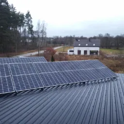 Instalacja fotowoltaiczna na dachu z blachy trapezowej, w tle dom jednorodzinny i maszt z polską flagą.