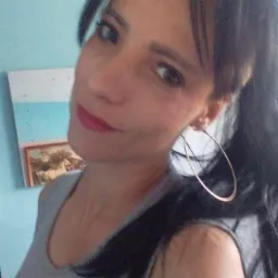 Portret kobiety z ciemnymi włosami, w szarej koszulce, z kolczykami kołami, na tle błękitnej ściany z obrazkiem i małym lustrem.