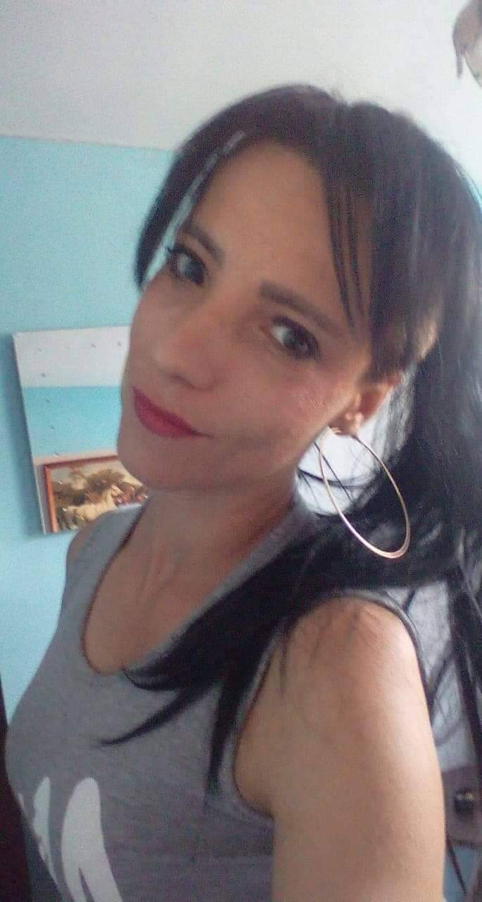 Portret kobiety z ciemnymi włosami, w szarej koszulce, z kolczykami kołami, na tle błękitnej ściany z obrazkiem i małym lustrem.