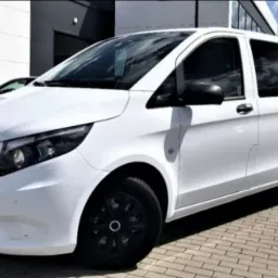 Biały Mercedes-Benz Vito z przyciemnianymi szybami, zaparkowany na kostce brukowej przed budynkiem z napisem 'RENT' w tle.