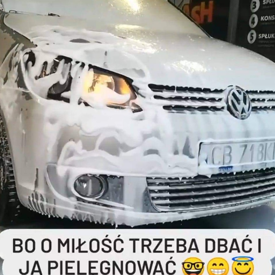 Przód srebrnego Volkswagena pokryty gęstą pianą podczas mycia w myjni samochodowej; widoczna tablica rejestracyjna i odblask w reflektorze.