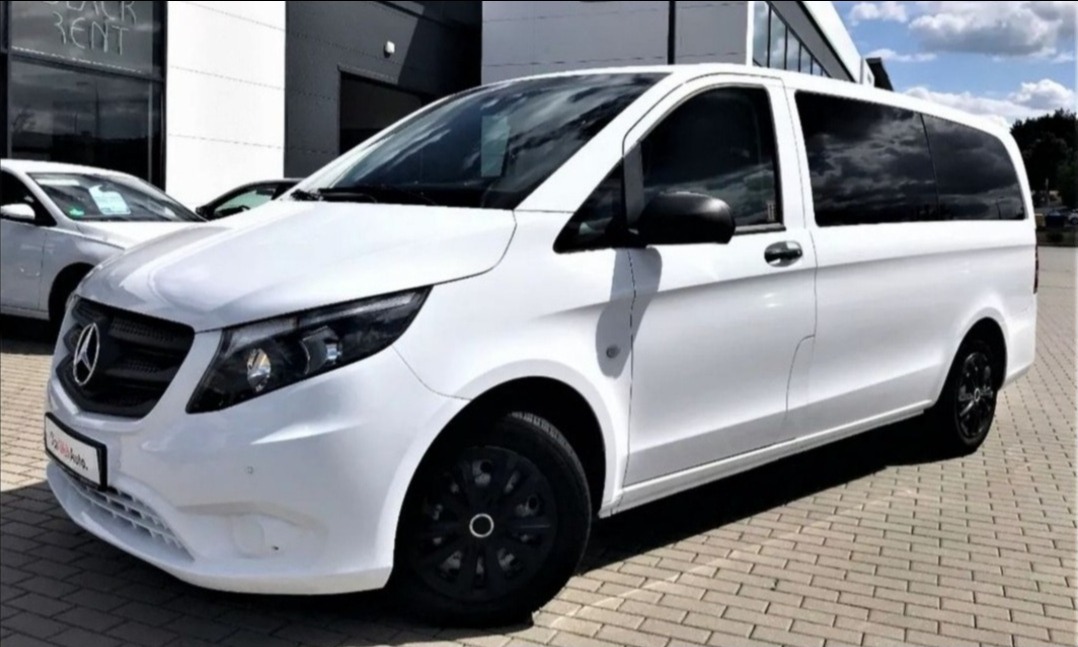 Biały Mercedes-Benz Vito z przyciemnianymi szybami, zaparkowany na kostce brukowej przed budynkiem z napisem 'RENT' w tle.