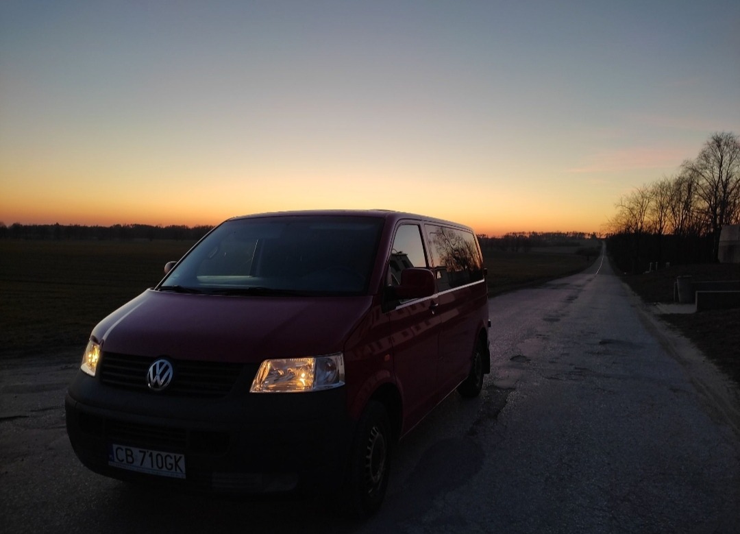 Czerwony Volkswagen Transporter T4 na wiejskiej drodze o zmierzchu, z włączonymi światłami i widoczną tablicą rejestracyjną CB 710GK.