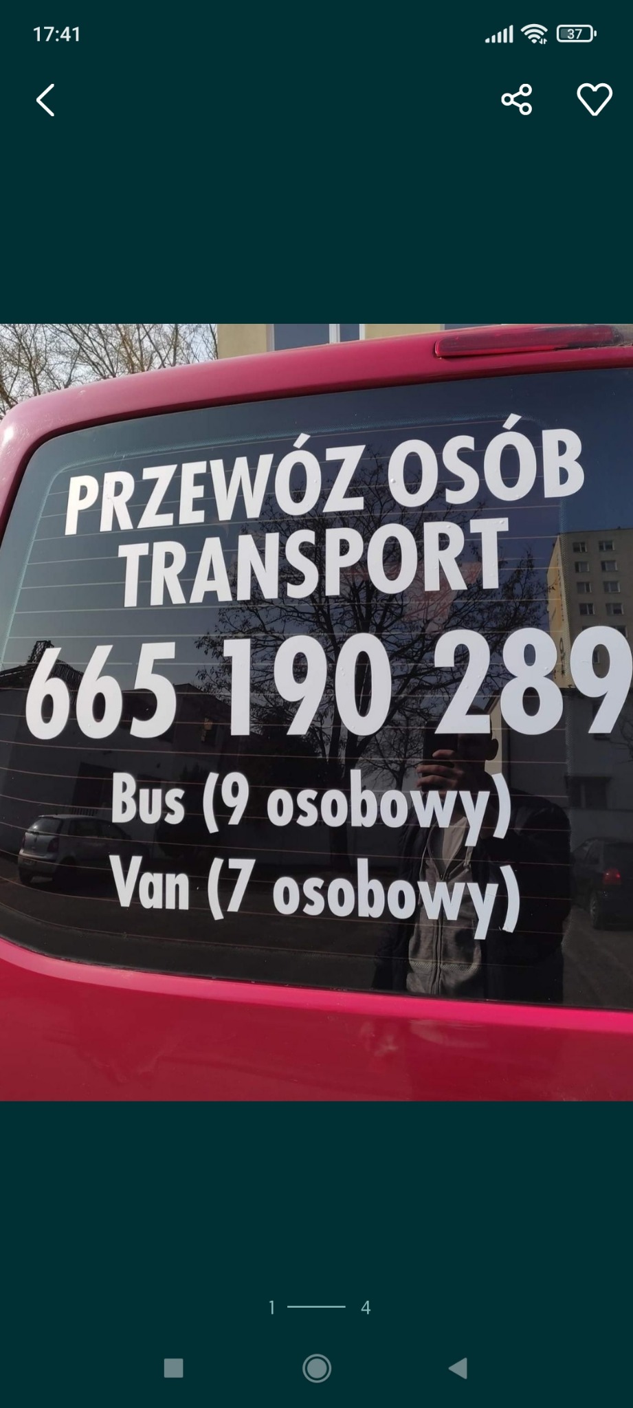 Tylna szyba czerwonego busa z białymi napisami informującymi o numerze telefonu i ofercie przewozu osób busem 9-osobowym i vanem 7-osobowym, odbicie fotografa w szybie.