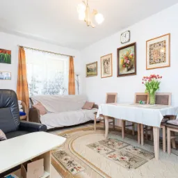 Jasny salon z czarnym skórzanym fotelem, beżową sofą przy oknie z pomarańczowymi zasłonami, stołem z białym obrusem i bukietem tulipanów, dywanem z motywem kwiatowym, oraz obrazami na ścianie.