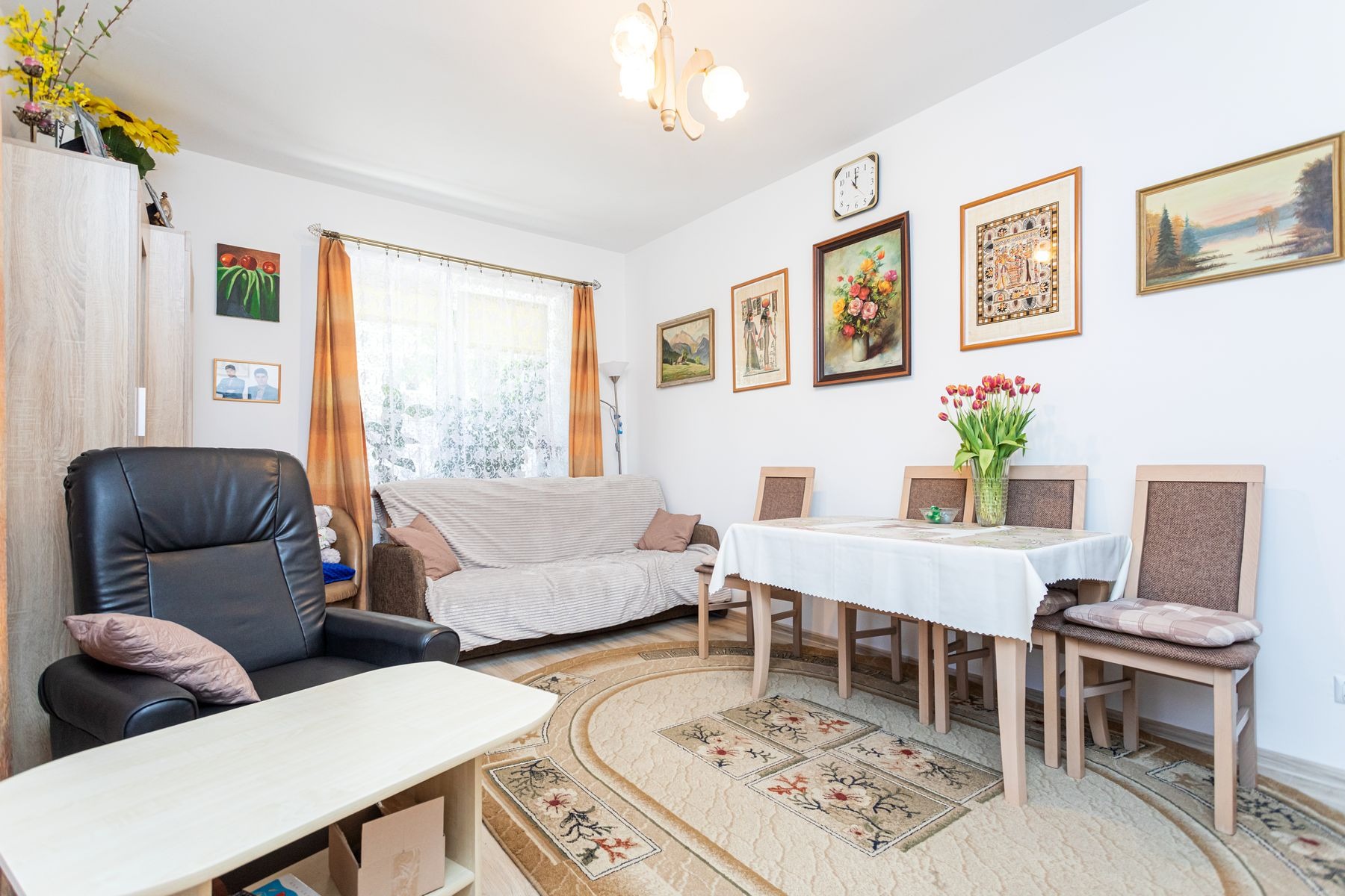 Jasny salon z czarnym skórzanym fotelem, beżową sofą przy oknie z pomarańczowymi zasłonami, stołem z białym obrusem i bukietem tulipanów, dywanem z motywem kwiatowym, oraz obrazami na ścianie.