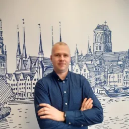 Mężczyzna w niebieskiej koszuli zapiętej na guziki, stojący na tle ściany z rysunkiem panoramy miasta w odcieniach niebieskiego.