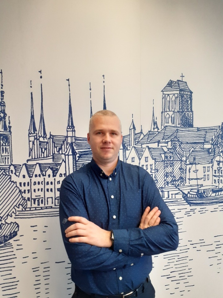 Mężczyzna w niebieskiej koszuli zapiętej na guziki, stojący na tle ściany z rysunkiem panoramy miasta w odcieniach niebieskiego.