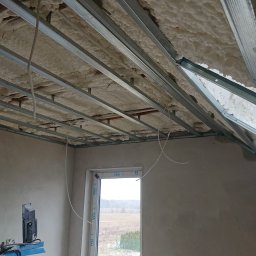 Robert Stachowiak StachBUD - Poddasze w trakcie izolacji, widoczne metalowe profile konstrukcyjne sufitu podwieszanego z wypełnieniem z pianki izolacyjnej, okno z widokiem na zimowy krajobraz, ściany w stanie surowym.